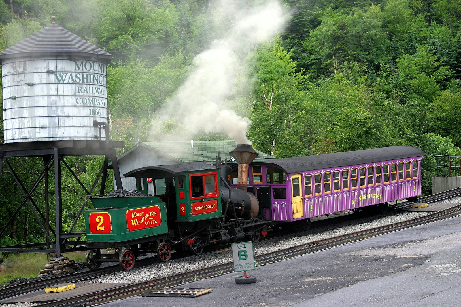 A green steam locomotive pushing a purple passenger coach beside a wooden water tower painted with the railway's name, smoke drifting into the forest