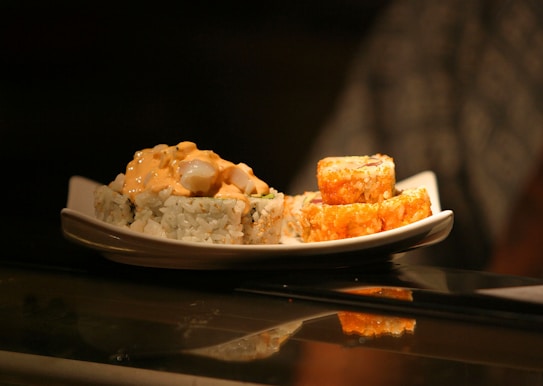 A plate of sushi rolls is presented on a reflective surface. The rolls are garnished with a creamy orange sauce and the lighting creates a warm, inviting atmosphere.
