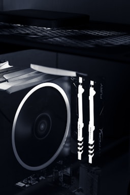 A close-up view of the interior of a computer case featuring illuminated RAM sticks with the brand name 'HyperX' visible. A large cooling fan is adjacent, showcasing the high-tech components of a gaming or performance PC setup.