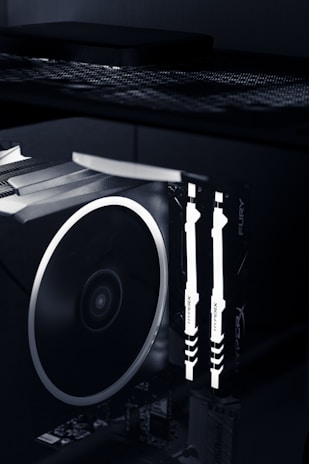 A close-up view of the interior of a computer case featuring illuminated RAM sticks with the brand name 'HyperX' visible. A large cooling fan is adjacent, showcasing the high-tech components of a gaming or performance PC setup.