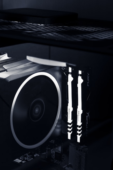 A close-up view of the interior of a computer case featuring illuminated RAM sticks with the brand name 'HyperX' visible. A large cooling fan is adjacent, showcasing the high-tech components of a gaming or performance PC setup.