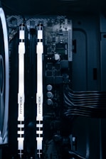 A detailed view of a computer's internal components featuring two illuminated HyperX RAM sticks, a cooling fan on the left, various capacitors, and electronic circuits on the motherboard. Cables are neatly organized on the right side.