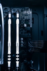 A detailed view of a computer's internal components featuring two illuminated HyperX RAM sticks, a cooling fan on the left, various capacitors, and electronic circuits on the motherboard. Cables are neatly organized on the right side.