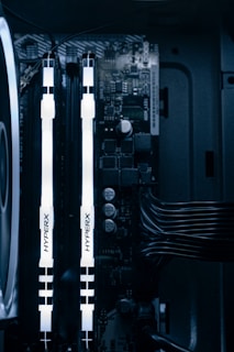 A detailed view of a computer's internal components featuring two illuminated HyperX RAM sticks, a cooling fan on the left, various capacitors, and electronic circuits on the motherboard. Cables are neatly organized on the right side.