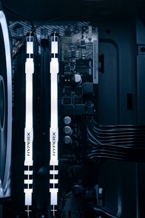 A detailed view of a computer's internal components featuring two illuminated HyperX RAM sticks, a cooling fan on the left, various capacitors, and electronic circuits on the motherboard. Cables are neatly organized on the right side.