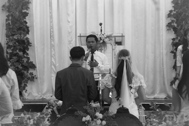 A black and white image of a wedding ceremony with a couple at the altar facing a minister. They are surrounded by floral decorations and guests. The bride is wearing a dress with lace details and a long veil.