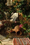 A cozy outdoor setting featuring lush greenery with a variety of plants. A large clay pot lies on the ground, next to a wooden bowl containing decorative items. A lantern hangs above within the branches. Colorful striped textiles are placed on the ground, with a striped cushion leaning against a white wooden bench. Pink flowers add a touch of color against the natural stone and green leaves.