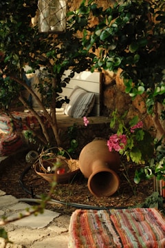 A cozy outdoor setting featuring lush greenery with a variety of plants. A large clay pot lies on the ground, next to a wooden bowl containing decorative items. A lantern hangs above within the branches. Colorful striped textiles are placed on the ground, with a striped cushion leaning against a white wooden bench. Pink flowers add a touch of color against the natural stone and green leaves.