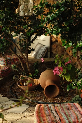 A cozy outdoor setting featuring lush greenery with a variety of plants. A large clay pot lies on the ground, next to a wooden bowl containing decorative items. A lantern hangs above within the branches. Colorful striped textiles are placed on the ground, with a striped cushion leaning against a white wooden bench. Pink flowers add a touch of color against the natural stone and green leaves.