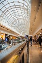 A brightly lit shopping mall features a spacious atrium with a large arched glass ceiling that allows natural light to flood the interior. People are walking and shopping, with various store fronts visible along the sides. An escalator in the foreground leads to a lower level, and the scene is lively and bustling.
