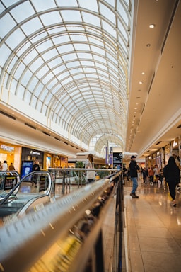 A brightly lit shopping mall features a spacious atrium with a large arched glass ceiling that allows natural light to flood the interior. People are walking and shopping, with various store fronts visible along the sides. An escalator in the foreground leads to a lower level, and the scene is lively and bustling.
