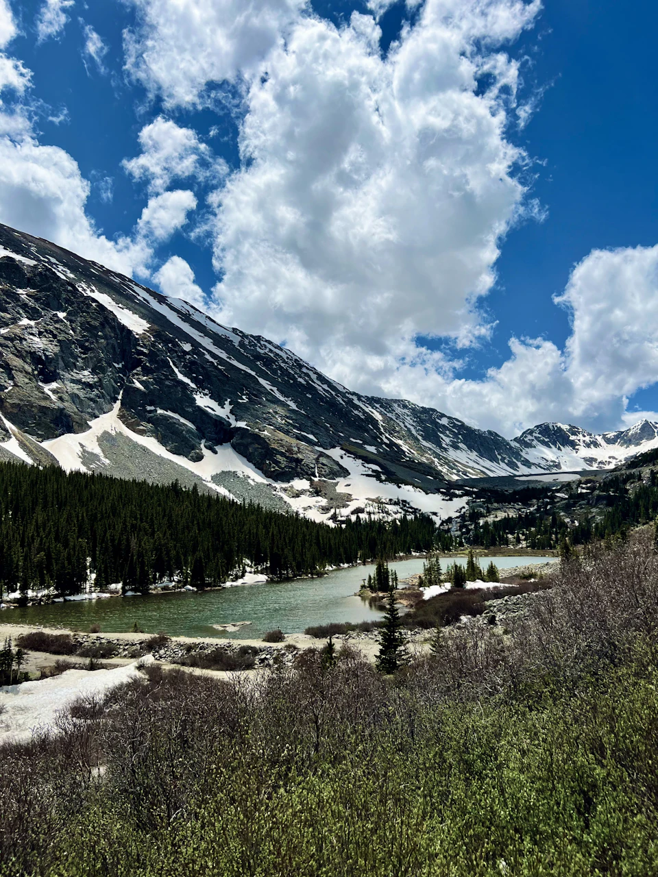 The image depicts a serene mountain landscape with snow-capped peaks and a deep blue sky dotted with fluffy white clouds. The mountains, covered in snow, stretch across the background, creating a sense of depth and grandeur. In the foreground, there is a small lake surrounded by lush green trees and bushes, adding a touch of life to the otherwise barren landscape. The image is taken from a high vantage point, providing a panoramic view of the mountains and their surroundings.