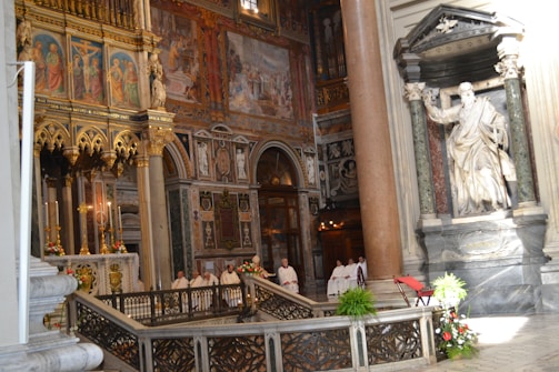 A richly decorated church interior features intricate religious artwork, statues, and ornate architectural details. There are clergy members in white robes standing near the altar, suggesting a ceremonial or religious event. The walls are adorned with detailed frescoes and gold embellishments.