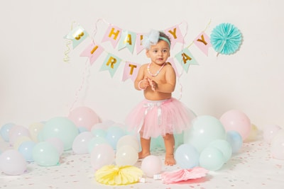 A young child is standing in a decorated room wearing a light pink tutu and a blue hair bow. The background is adorned with a 'Happy Birthday' banner and pastel-colored balloons. The child is surrounded by various colorful balloons and paper decorations on the floor.