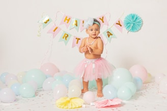 A young child is standing in a decorated room wearing a light pink tutu and a blue hair bow. The background is adorned with a 'Happy Birthday' banner and pastel-colored balloons. The child is surrounded by various colorful balloons and paper decorations on the floor.