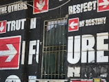 A wall is covered with bold, large text and arrows on a dark background. Words such as 'TRUTH,' 'FUTURE,' 'RESPECT,' and 'DESTINY' are prominently displayed in white with red arrow symbols pointing in various directions. There is a barred window with some fabric draping inside, which adds an urban, gritty feel to the scene.