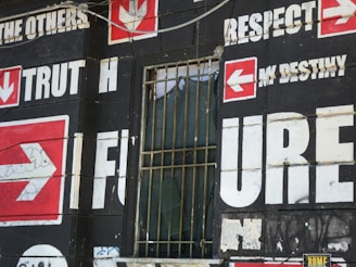 A wall is covered with bold, large text and arrows on a dark background. Words such as 'TRUTH,' 'FUTURE,' 'RESPECT,' and 'DESTINY' are prominently displayed in white with red arrow symbols pointing in various directions. There is a barred window with some fabric draping inside, which adds an urban, gritty feel to the scene.