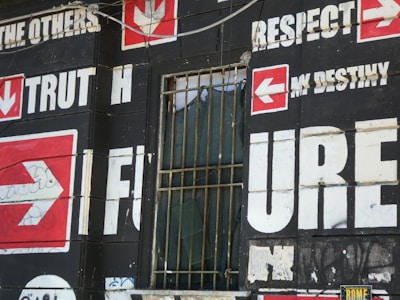 A wall is covered with bold, large text and arrows on a dark background. Words such as 'TRUTH,' 'FUTURE,' 'RESPECT,' and 'DESTINY' are prominently displayed in white with red arrow symbols pointing in various directions. There is a barred window with some fabric draping inside, which adds an urban, gritty feel to the scene.