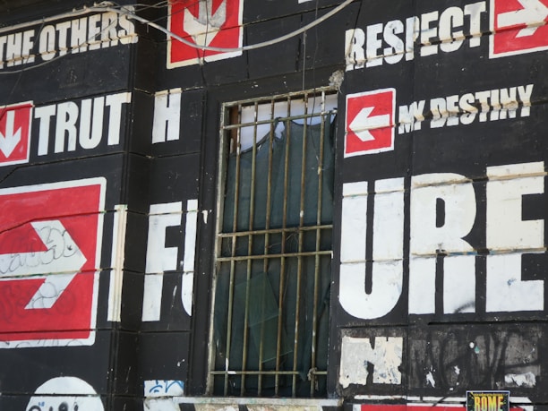 A wall is covered with bold, large text and arrows on a dark background. Words such as 'TRUTH,' 'FUTURE,' 'RESPECT,' and 'DESTINY' are prominently displayed in white with red arrow symbols pointing in various directions. There is a barred window with some fabric draping inside, which adds an urban, gritty feel to the scene.