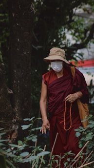 A person dressed in a maroon robe stands in a lush, green forested area. They are wearing a beige sun hat and a white face mask. In one hand, they hold a string of orange prayer beads. The setting is serene, with a mixture of natural foliage surrounding them.