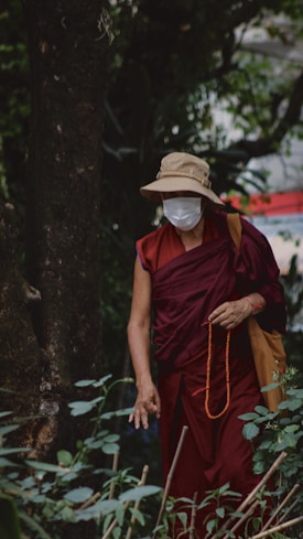 A person dressed in a maroon robe stands in a lush, green forested area. They are wearing a beige sun hat and a white face mask. In one hand, they hold a string of orange prayer beads. The setting is serene, with a mixture of natural foliage surrounding them.