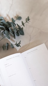 An open planner is displayed on a light marble surface, with a sprig of eucalyptus leaves placed on the table next to it. The planner shows sections for weekly and daily planning.