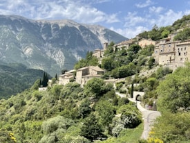 A picturesque hillside village with stone buildings nestled among lush green vegetation. The houses are set on a slope with a backdrop of tall, rugged mountains and a partially cloudy sky. A winding road leads up through the village, with cypress trees lining the path.