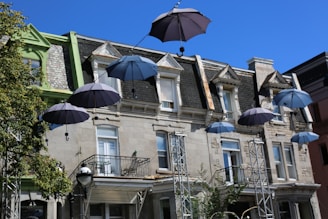 Several open umbrellas are suspended in the air in front of an old stone building with a steep roof and decorative architecture. The umbrellas are attached to wires, creating a whimsical display. The building has multiple windows with some balconies, and there is a tree and metal structures supporting the wires.