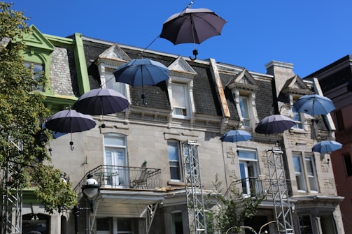 Several open umbrellas are suspended in the air in front of an old stone building with a steep roof and decorative architecture. The umbrellas are attached to wires, creating a whimsical display. The building has multiple windows with some balconies, and there is a tree and metal structures supporting the wires.
