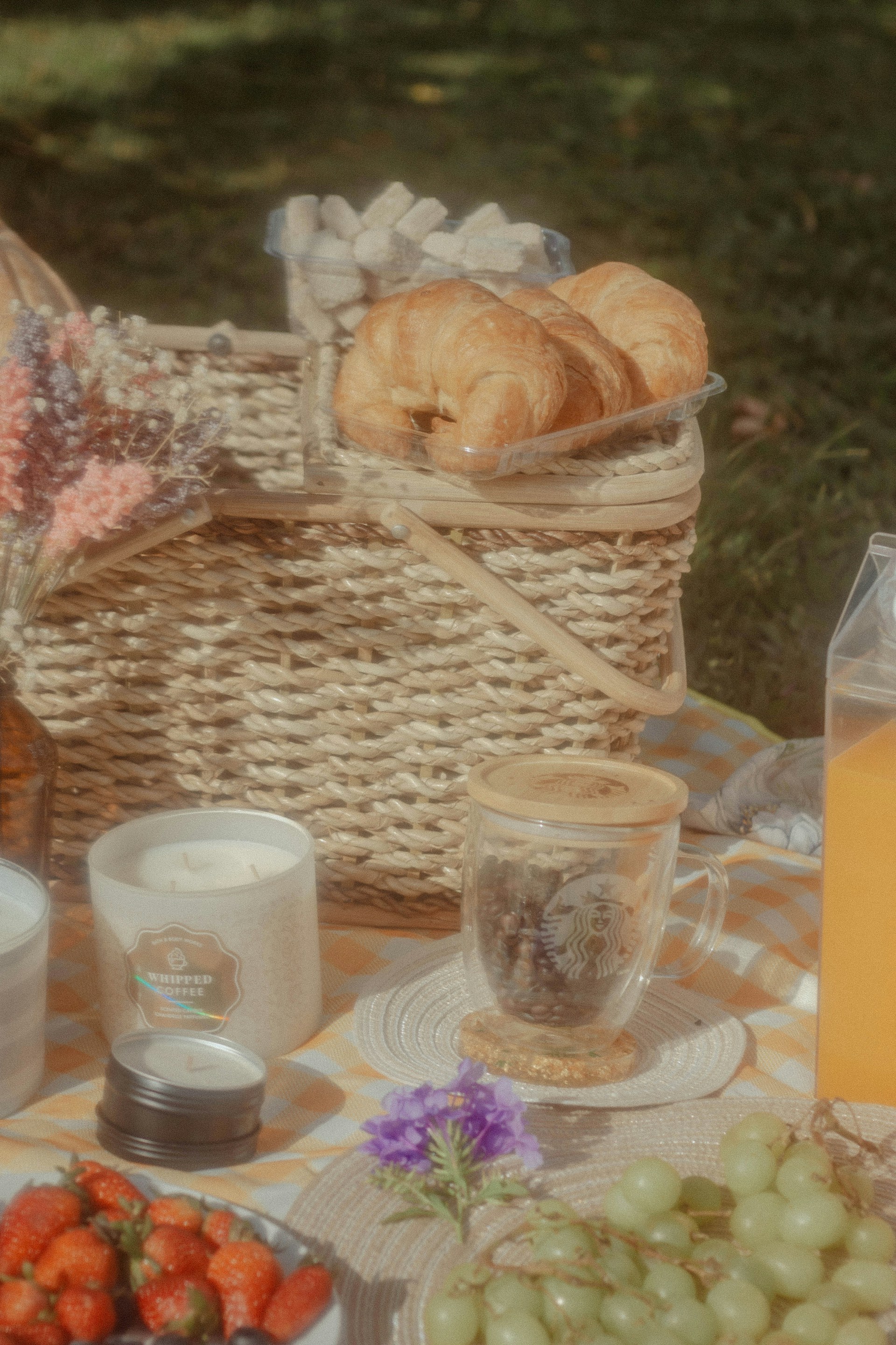 A picnic setup on a checkered cloth featuring a wicker basket filled with croissants and sugar cubes. Nearby, there's a jar of whipped coffee, a glass cup with coffee beans, a candle, and plates with strawberries and green grapes. Bright flowers add a bit of color to the scene.