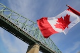 A large Canadian flag waves prominently in the sky, with a green steel bridge structure stretching diagonally across the background. The sky is mostly clear with a few wispy clouds.