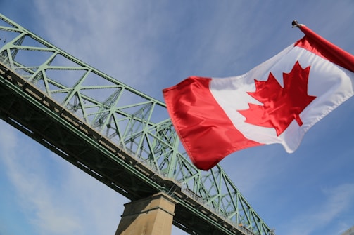 A large Canadian flag waves prominently in the sky, with a green steel bridge structure stretching diagonally across the background. The sky is mostly clear with a few wispy clouds.