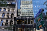 A juxtaposition of architectural styles featuring a historic building with arch windows next to a modern glass skyscraper. Reflective surfaces create interesting visual effects. Street signs for 'Square Victoria' and 'Cité Internationale' indicate the location, and there are hints of greenery with visible tree branches.