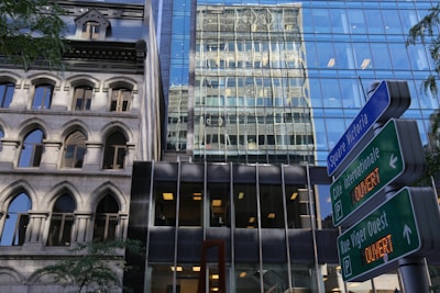 A juxtaposition of architectural styles featuring a historic building with arch windows next to a modern glass skyscraper. Reflective surfaces create interesting visual effects. Street signs for 'Square Victoria' and 'Cité Internationale' indicate the location, and there are hints of greenery with visible tree branches.