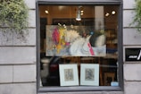 A storefront window displays a modern abstract painting featuring a stylized horse with vibrant splashes of color such as yellow, pink, and red. Below the painting are two framed sketches or prints. The window is flanked by gray stone walls with greenery hanging on each side, adding a touch of nature to the urban setting. Inside, warm light illuminates the art, providing an inviting appearance.