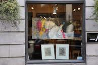 A storefront window displays a modern abstract painting featuring a stylized horse with vibrant splashes of color such as yellow, pink, and red. Below the painting are two framed sketches or prints. The window is flanked by gray stone walls with greenery hanging on each side, adding a touch of nature to the urban setting. Inside, warm light illuminates the art, providing an inviting appearance.