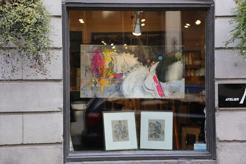 A storefront window displays a modern abstract painting featuring a stylized horse with vibrant splashes of color such as yellow, pink, and red. Below the painting are two framed sketches or prints. The window is flanked by gray stone walls with greenery hanging on each side, adding a touch of nature to the urban setting. Inside, warm light illuminates the art, providing an inviting appearance.
