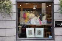 A storefront window displays a modern abstract painting featuring a stylized horse with vibrant splashes of color such as yellow, pink, and red. Below the painting are two framed sketches or prints. The window is flanked by gray stone walls with greenery hanging on each side, adding a touch of nature to the urban setting. Inside, warm light illuminates the art, providing an inviting appearance.