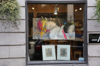 A storefront window displays a modern abstract painting featuring a stylized horse with vibrant splashes of color such as yellow, pink, and red. Below the painting are two framed sketches or prints. The window is flanked by gray stone walls with greenery hanging on each side, adding a touch of nature to the urban setting. Inside, warm light illuminates the art, providing an inviting appearance.