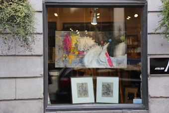 A storefront window displays a modern abstract painting featuring a stylized horse with vibrant splashes of color such as yellow, pink, and red. Below the painting are two framed sketches or prints. The window is flanked by gray stone walls with greenery hanging on each side, adding a touch of nature to the urban setting. Inside, warm light illuminates the art, providing an inviting appearance.