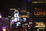 Bright city lights and large illuminated signs dominate a bustling urban skyline. Numerous advertisements and marquees for famous hotels and casinos are visible, showcasing entertainment and attractions in a vibrant nightlife setting.