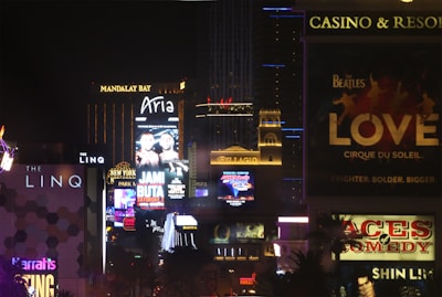 Bright city lights and large illuminated signs dominate a bustling urban skyline. Numerous advertisements and marquees for famous hotels and casinos are visible, showcasing entertainment and attractions in a vibrant nightlife setting.