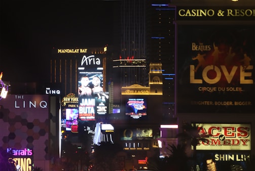 Bright city lights and large illuminated signs dominate a bustling urban skyline. Numerous advertisements and marquees for famous hotels and casinos are visible, showcasing entertainment and attractions in a vibrant nightlife setting.
