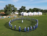 A group of people in blue uniforms and white helmets are standing in a circle on a grassy field. In the background, there are several white tents with logos and some trees. The sky is mostly clear, contributing to a bright and open atmosphere.