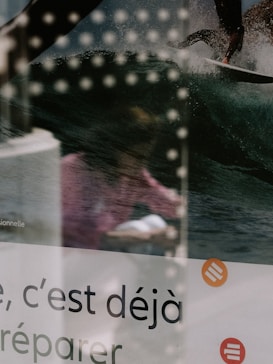 The image features a reflection of a person wearing a pink garment, visible on the glass surface. There is an overlay of a photograph showing a surfer on a wave, with water droplets frozen in motion. The lower part of the image contains French text along with logos or symbols in orange.