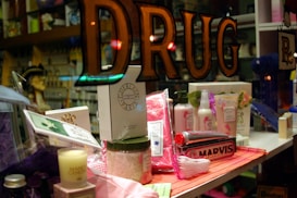 A shop window display featuring various toiletries and personal care products. Items include boxed perfumes, a candle, bath salts, and various bottles of lotions and creams. The presence of a dentist's tooth model and toothpaste at the forefront adds a focus on dental care. The word 'DRUG' is prominently displayed on the glass, suggesting a pharmacy or apothecary setting.