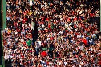 A densely packed crowd of people sitting in a stadium, dressed in casual summer clothing. Many are wearing hats and sunglasses, some are standing, and the atmosphere appears lively.