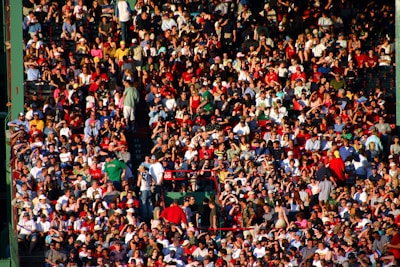 A densely packed crowd of people sitting in a stadium, dressed in casual summer clothing. Many are wearing hats and sunglasses, some are standing, and the atmosphere appears lively.