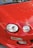 The close-up image features the front part of a red car with distinct circular headlights and smooth body contours. Reflections of trees are visible on the shiny surface of the car's hood, indicating that the car is outdoors.