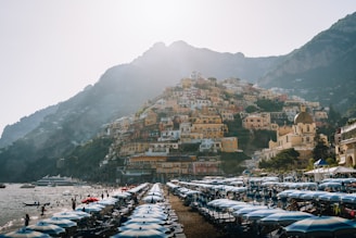 A picturesque coastal town with colorful hillside buildings overlooking a busy beach filled with sunbeds and umbrellas. The serene sea with boats and swimmers adds to the lively yet tranquil scene. The architecture features traditional Mediterranean design, characterized by vibrant facades and a domed church.